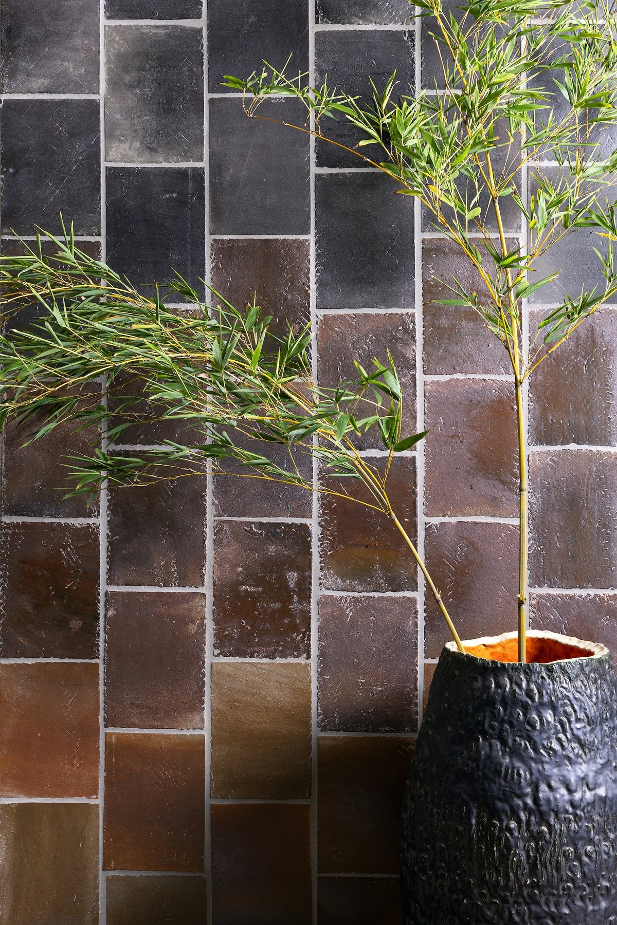 A wall of rectangular glazed tile laid in a vertical running bond, charcoal Kuro at the top grading through brown Kogecha to copper Suna at the bottom, with bamboo rising from a dark textured vessel