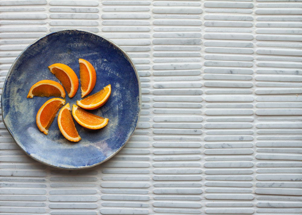 A blue glazed plate of orange segments arranged as a pinwheel, on a pale Carrara mosaic of narrow rounded sticks, seen from directly above